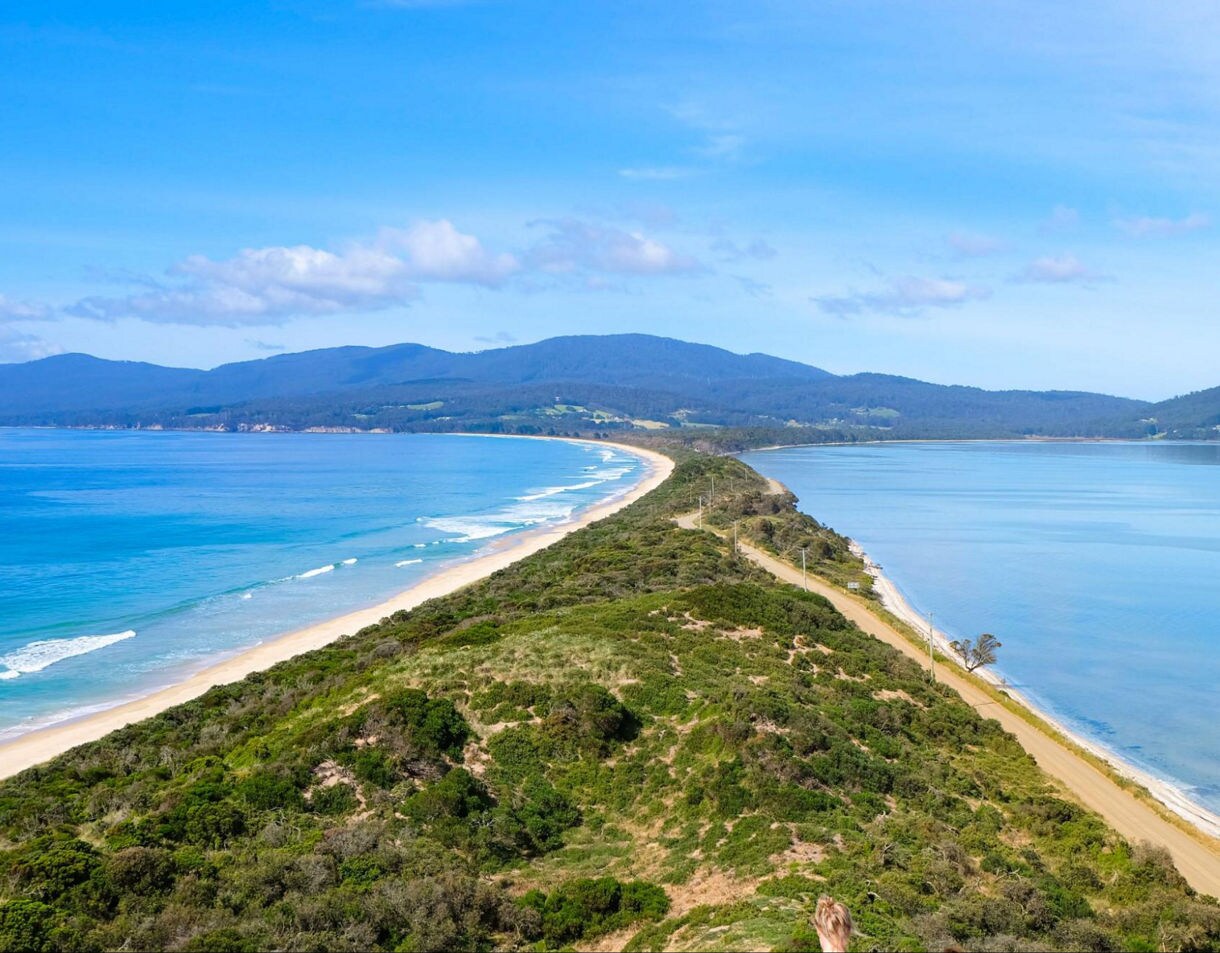 Aerial view of Bruny Island Neck in Tasmania, a narrow sandy isthmus with ocean on one side and calm bay waters on the other.