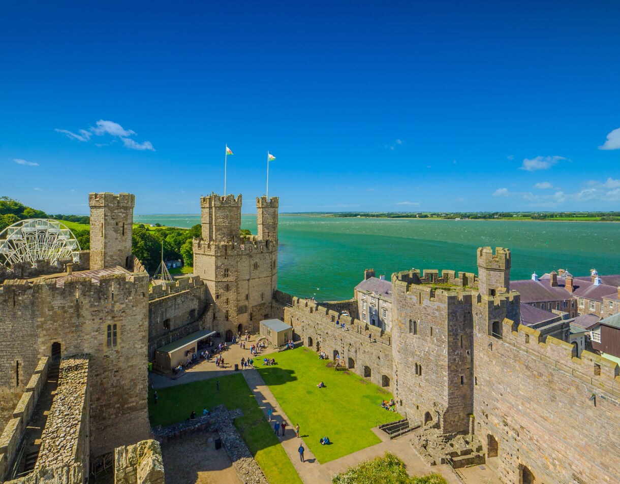 Aerial view of a medieval stone castle with towers and inner courtyard overlooking green lawns rooftops and a blue coastal inlet.