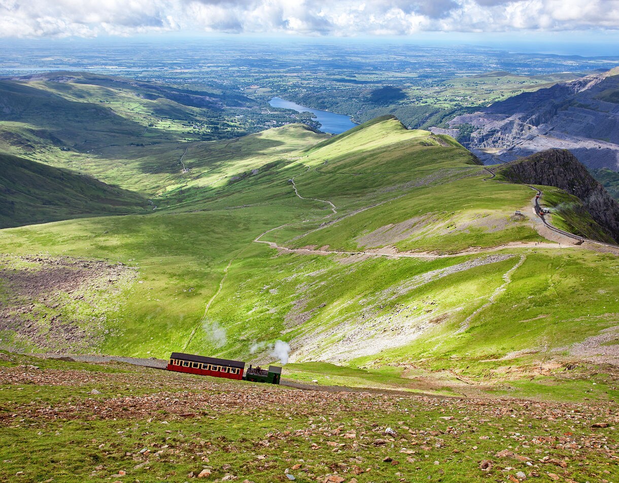 Scenic view of a small red train climbing a mountainside railway with rolling green hills valleys and distant lakes under a partly cloudy sky.