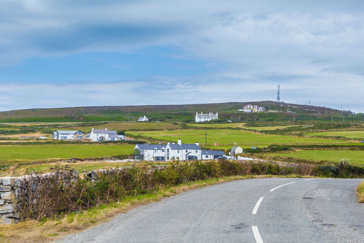 Curving rural road leading toward scattered white houses across rolling green fields under a soft blue sky.