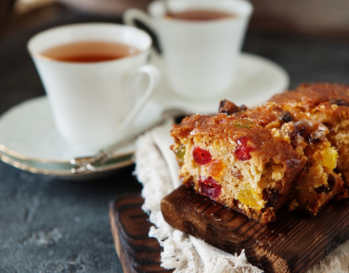 Close-up of sliced fruit loaf on a wooden board with two white cups of tea set on saucers in the background.