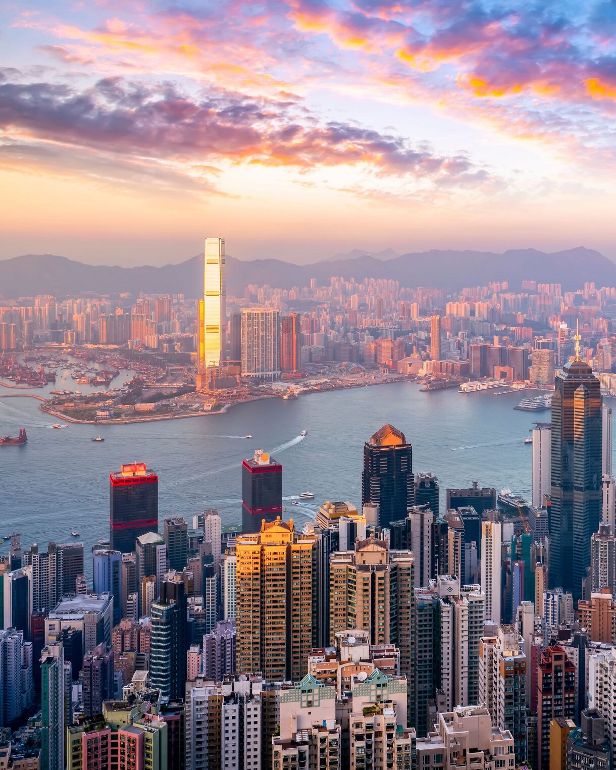 Panoramic view of Hong Kong’s skyline and Victoria Harbour at sunset, with glowing skyscrapers and colorful clouds above the city.