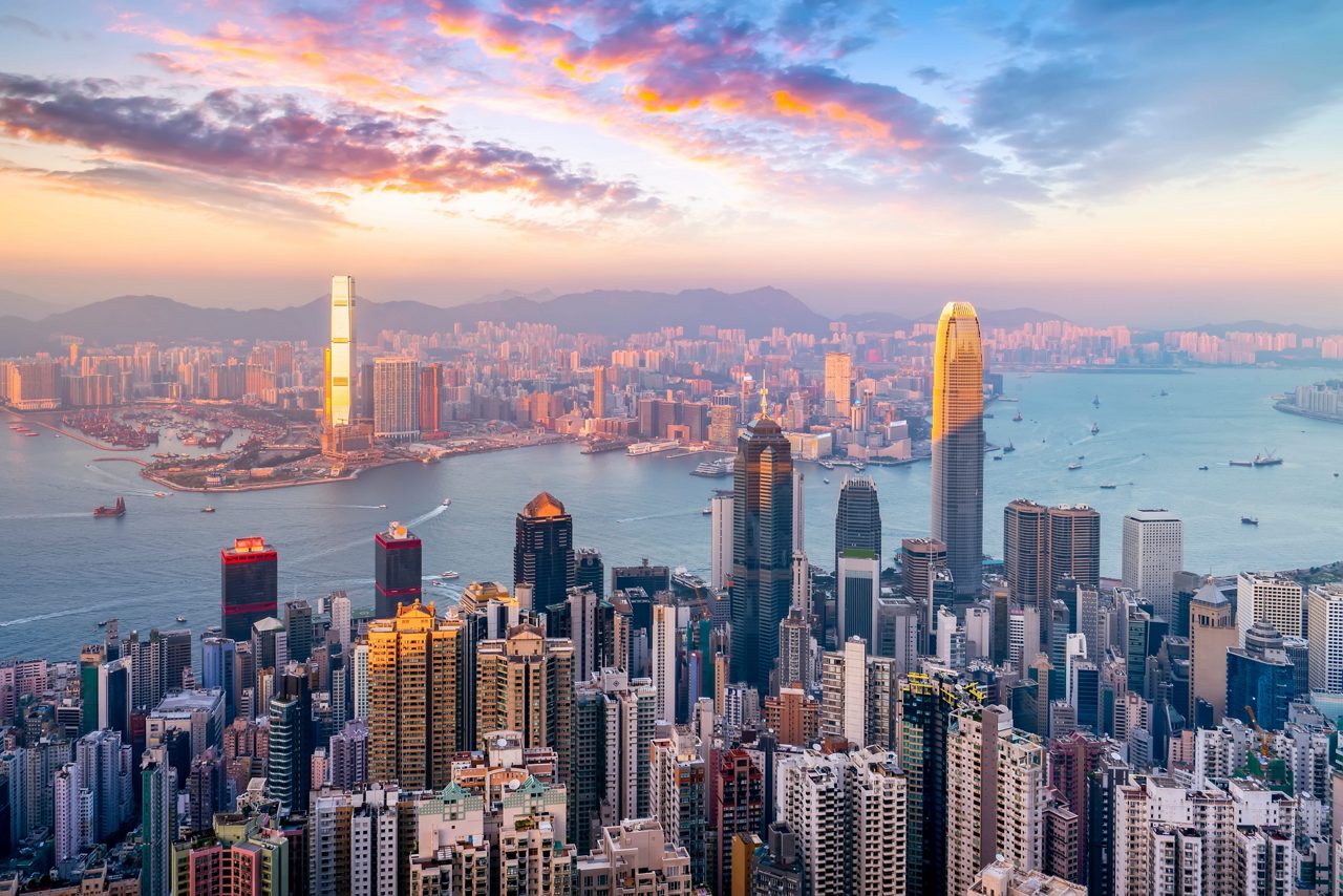 Panoramic view of Hong Kong’s skyline and Victoria Harbour at sunset, with glowing skyscrapers and colorful clouds above the city.