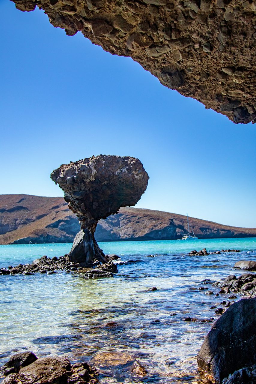 Mushroom-shaped rock formation standing in shallow clear water with desert hills and a sailboat in the background.