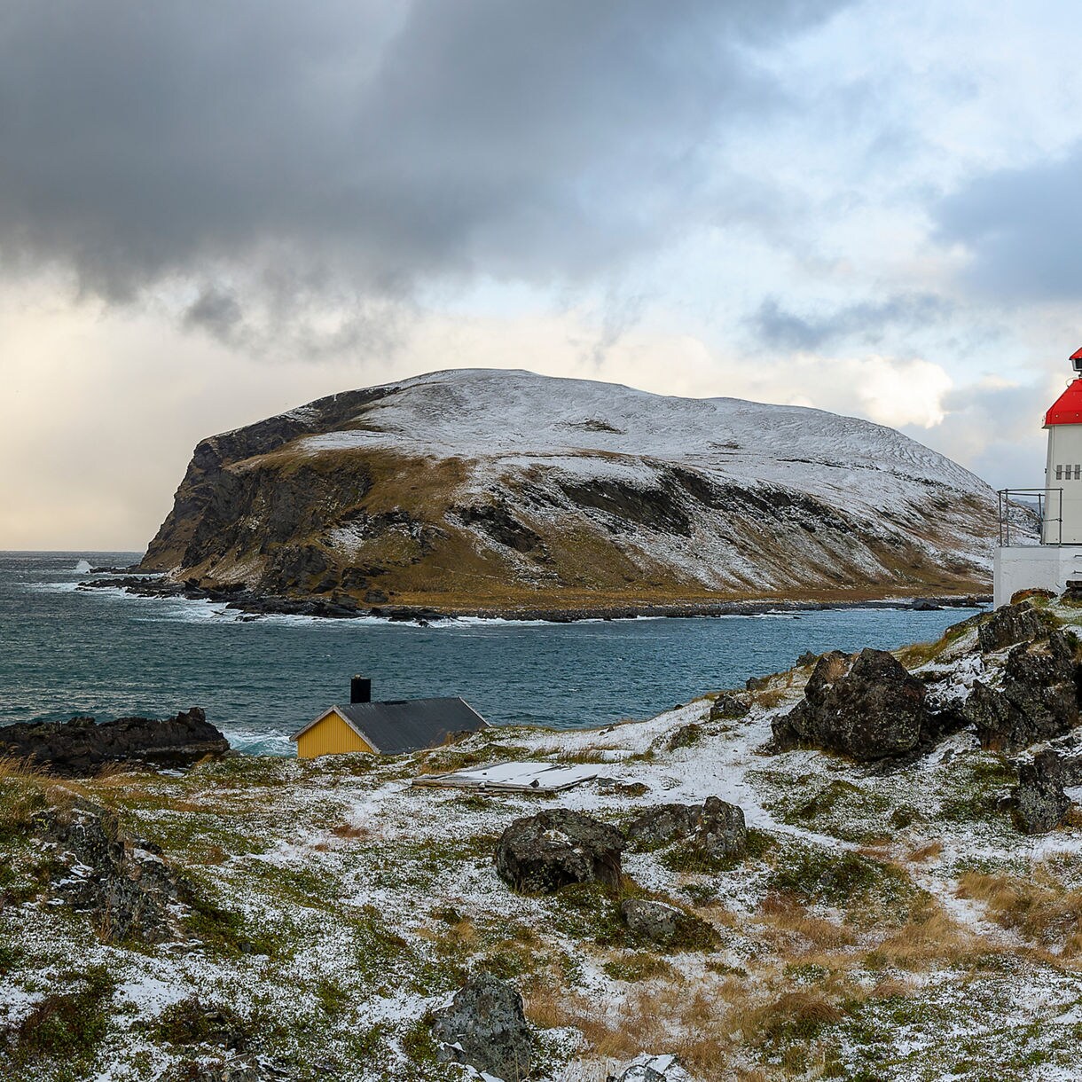 White lighthouse with a red top standing on rocky, snow-dusted terrain overlooking turquoise Arctic waters and distant islands under dramatic skies.