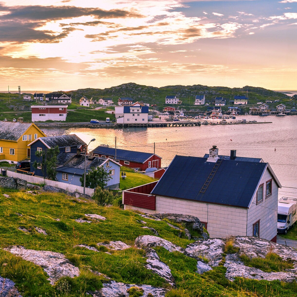 Panorama of Gjesvær, Norway, at sunset, with brightly painted houses, rocky shoreline and calm waters dotted with islands in the background.