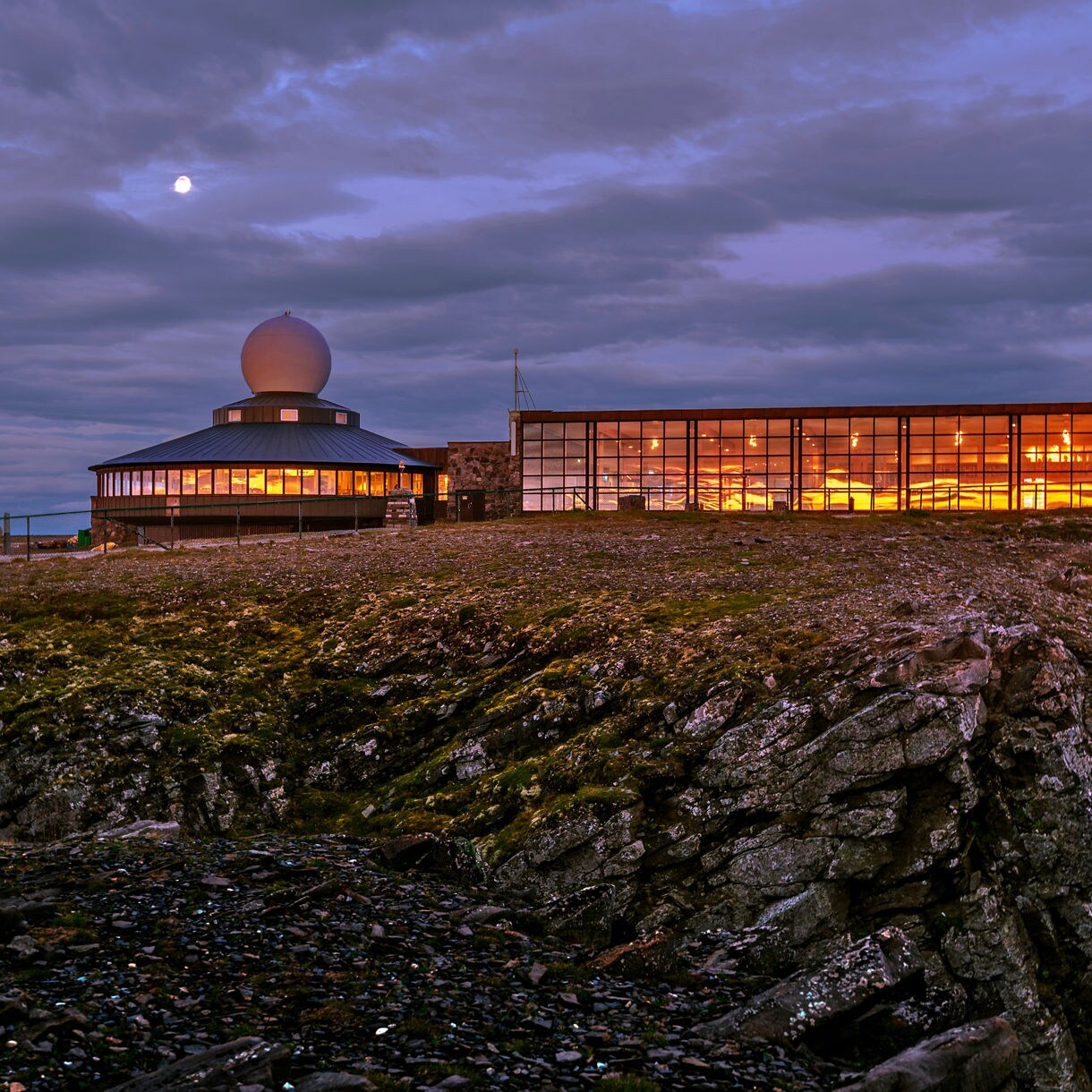 Nordkapp Hall in Norway at dusk, with warm lights glowing inside the glass-fronted building under a dramatic cloudy sky.