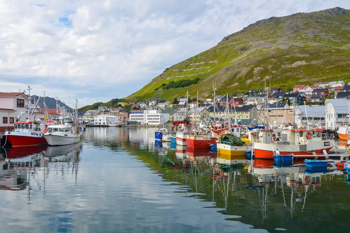 Fishing boats painted in bright reds, yellows and whites docked in Honningsvåg harbor, with hillside homes and green mountains rising in the background.