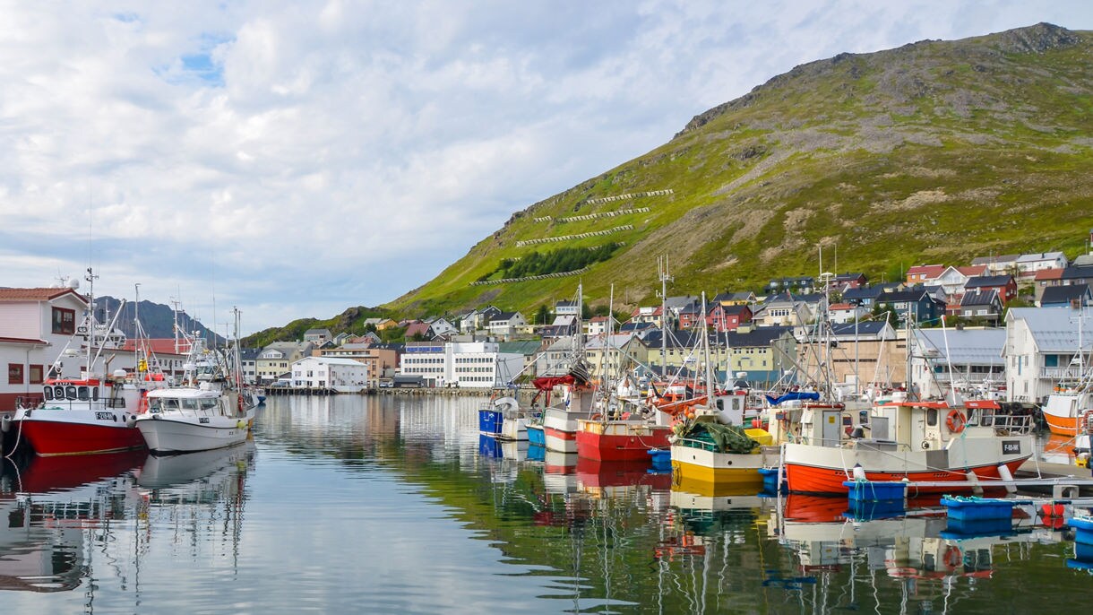 Fishing boats painted in bright reds, yellows and whites docked in Honningsvåg harbor, with hillside homes and green mountains rising in the background.