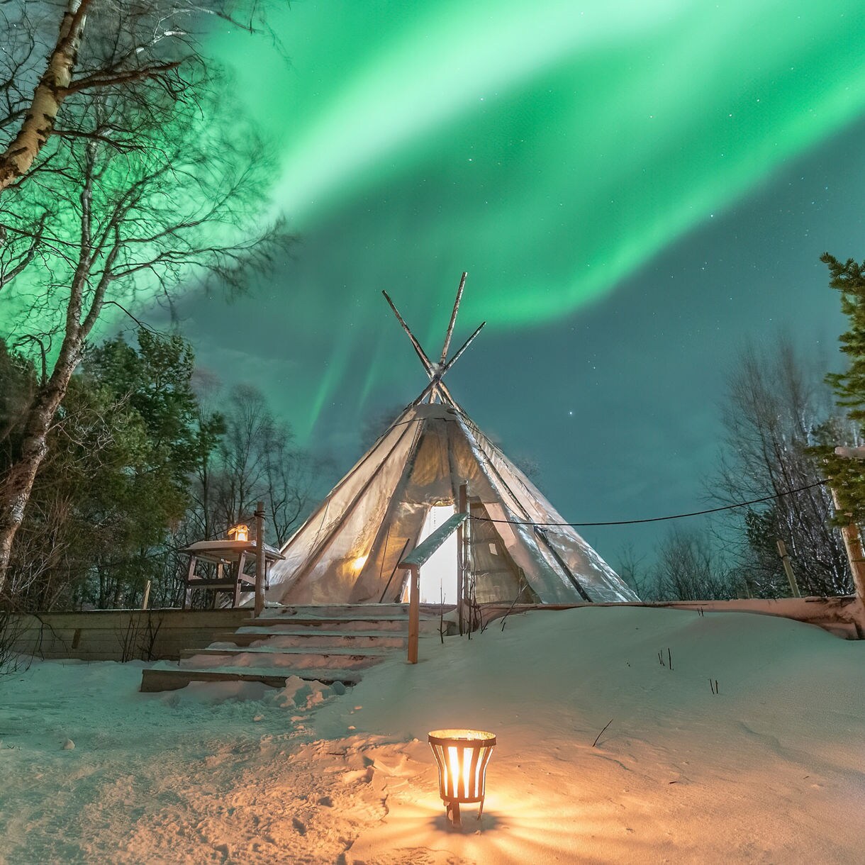 Traditional Sami lavvo tent glowing in snowy forest with vivid green aurora borealis streaking across the night sky.