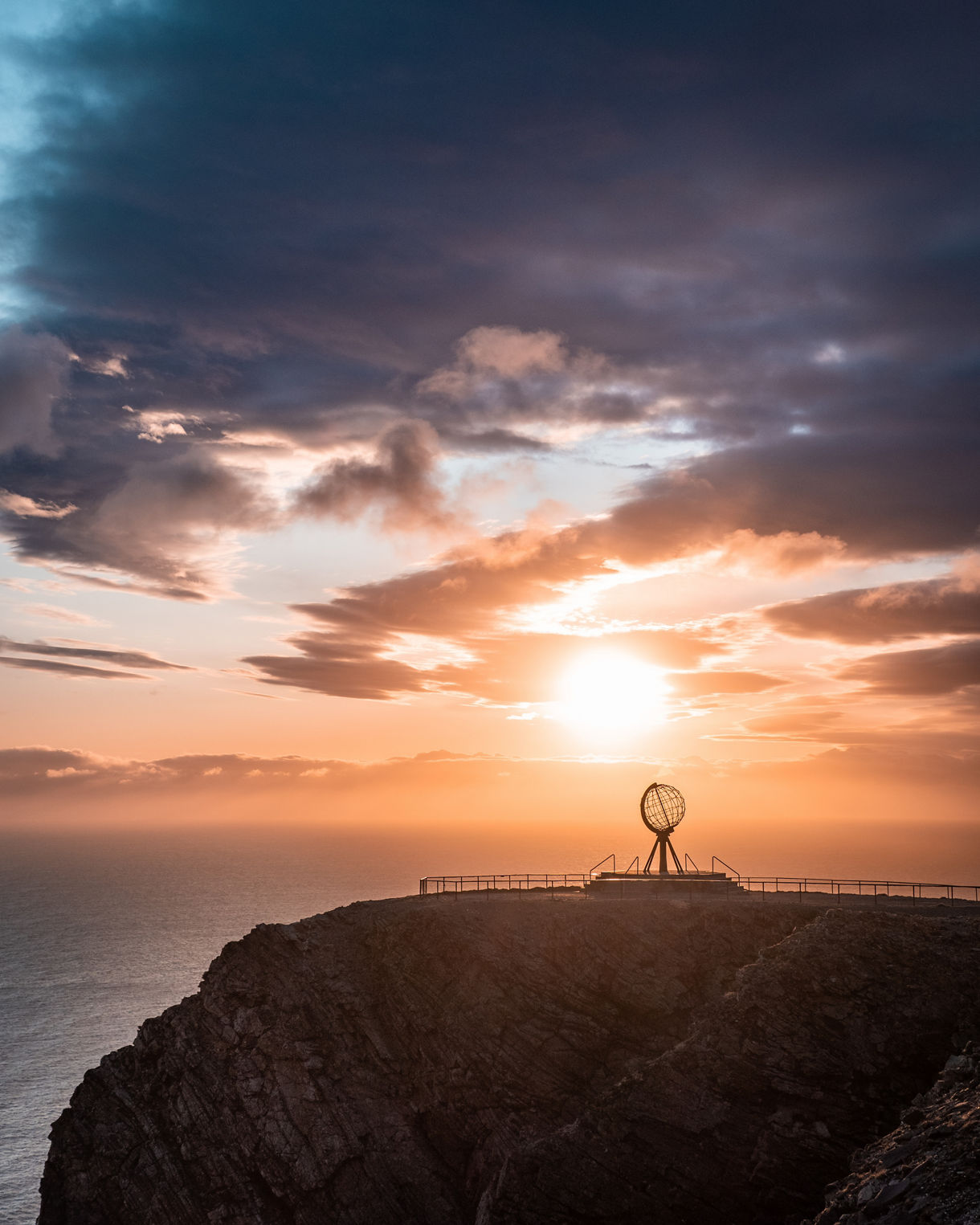 Silhouette of the North Cape Globe monument on a cliff at sunset with golden light over the Arctic Ocean.