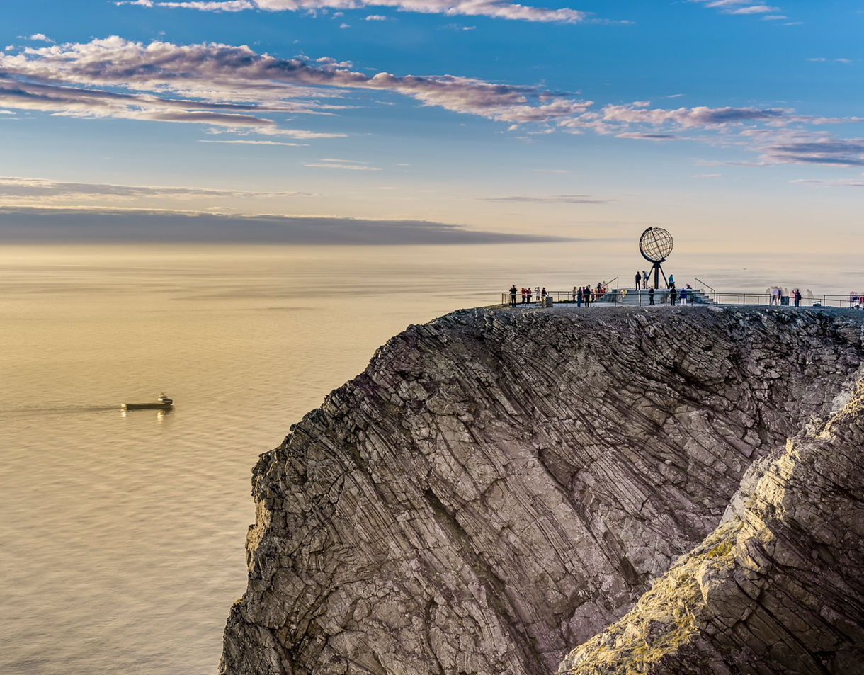 Clifftop viewpoint at the North Cape with the iconic globe monument, overlooking the golden Arctic Ocean with a small ship sailing in the distance.