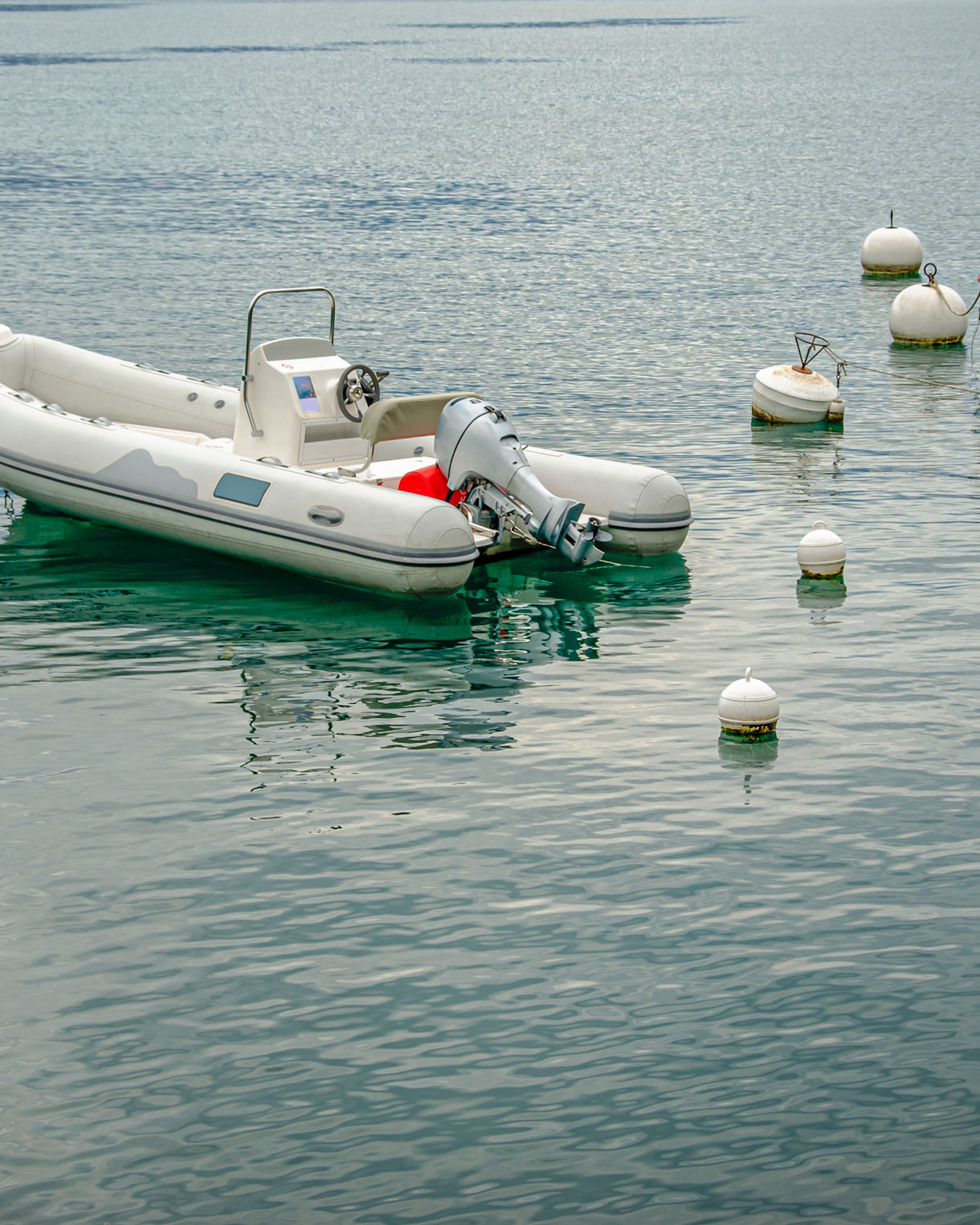 White inflatable motorboat floating among buoys on still, blue-green water with another small red boat nearby.