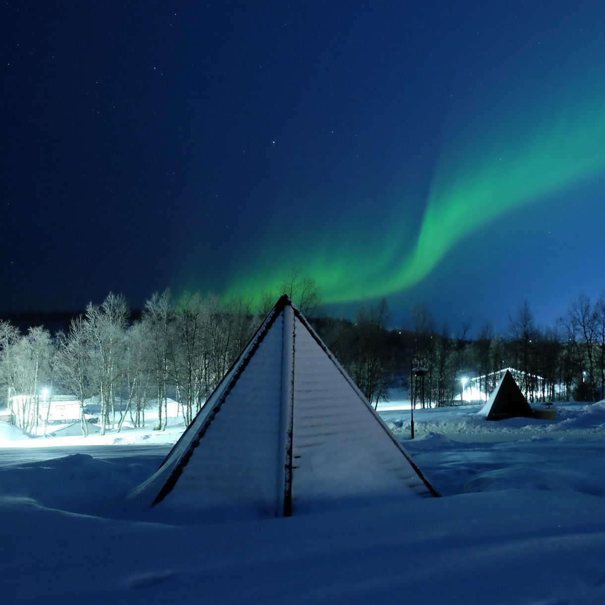 Green aurora borealis streaking across a starry sky above snow-covered Sámi-style tents and trees in a winter landscape.