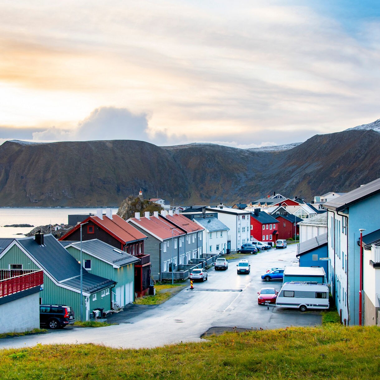 Colorful houses in Havøysund, Norway, with a small lighthouse in the distance, set against steep mountains and an Arctic coastline.