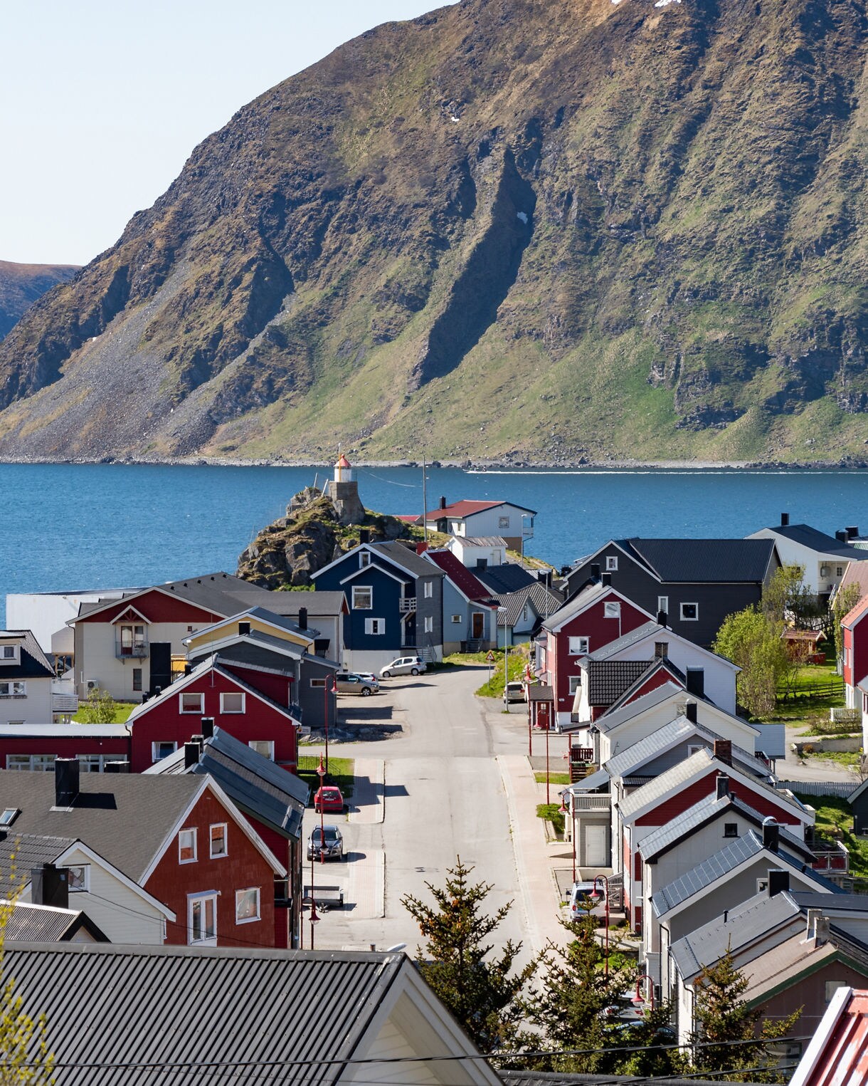 View of Honningsvåg, Norway, showing rows of red and gray houses leading toward a small lighthouse by the sea, with a towering green mountain rising across the fjord.