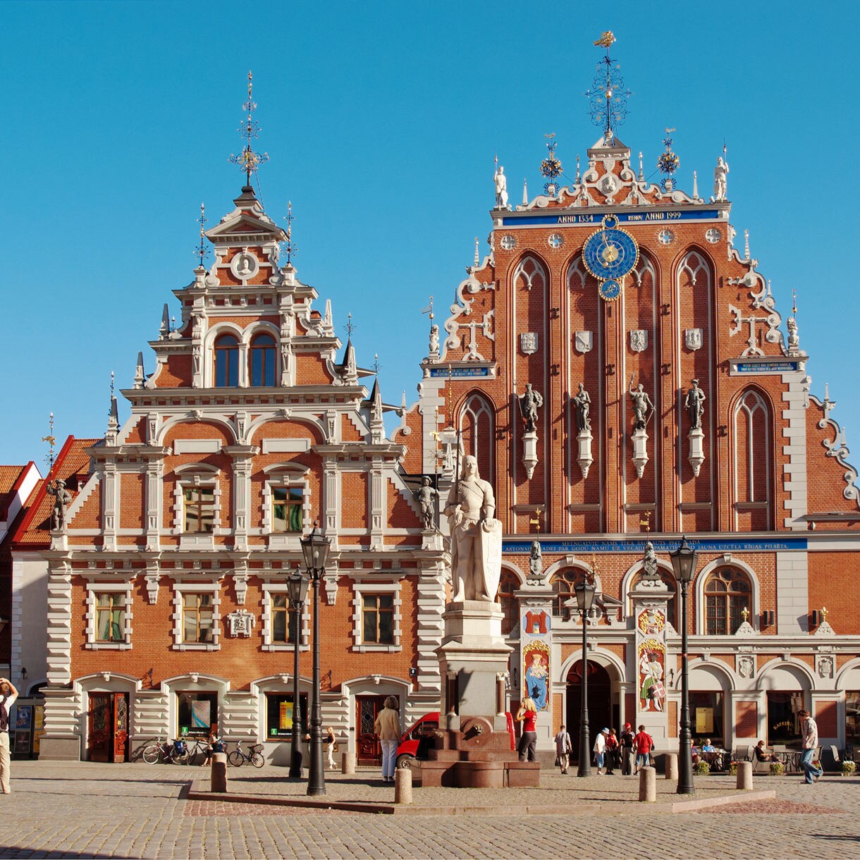Front view of the House of the Blackheads in Riga, Latvia, with its ornate red-brick façade, blue clock detail, white stone statues and richly decorated gables under a clear blue sky.
