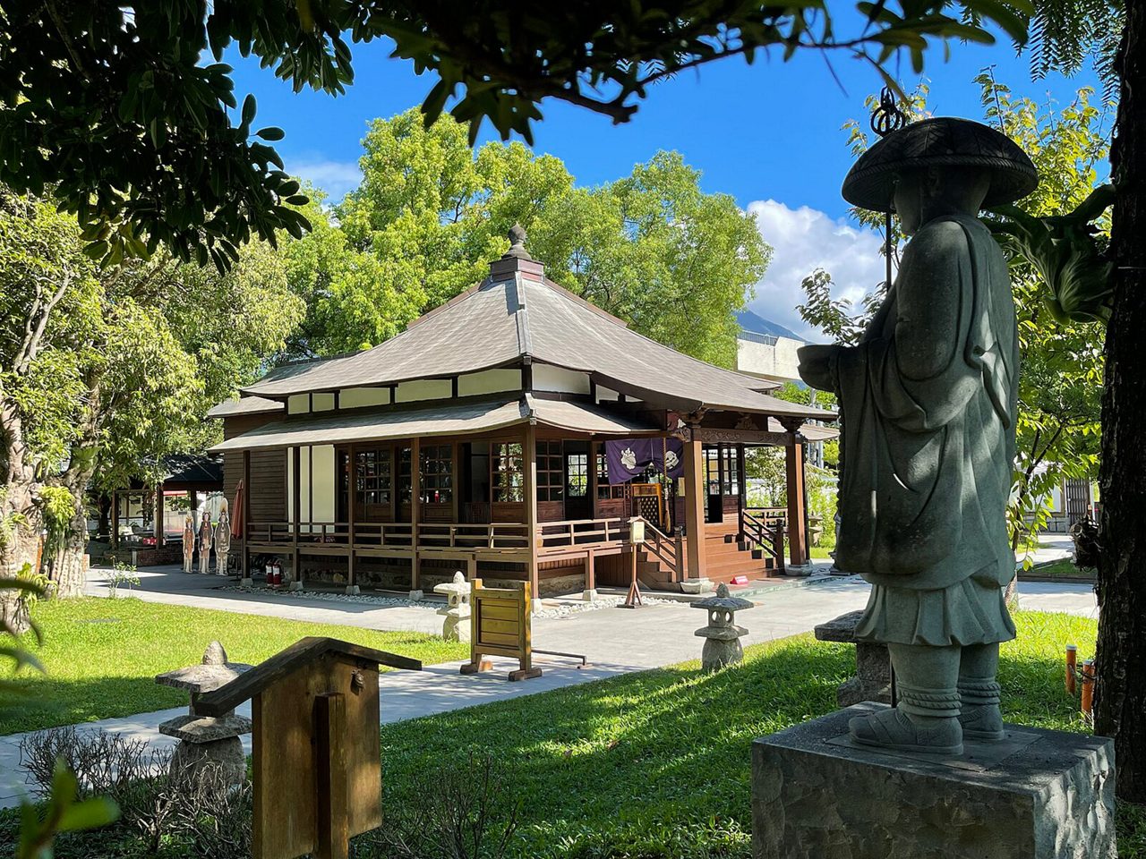 A tranquil Japanese-style temple in Hualien, Taiwan, featuring wooden architecture, a gently sloped roof, manicured lawns and stone lanterns. A statue of a robed figure wearing a hat stands in the foreground beneath leafy trees.