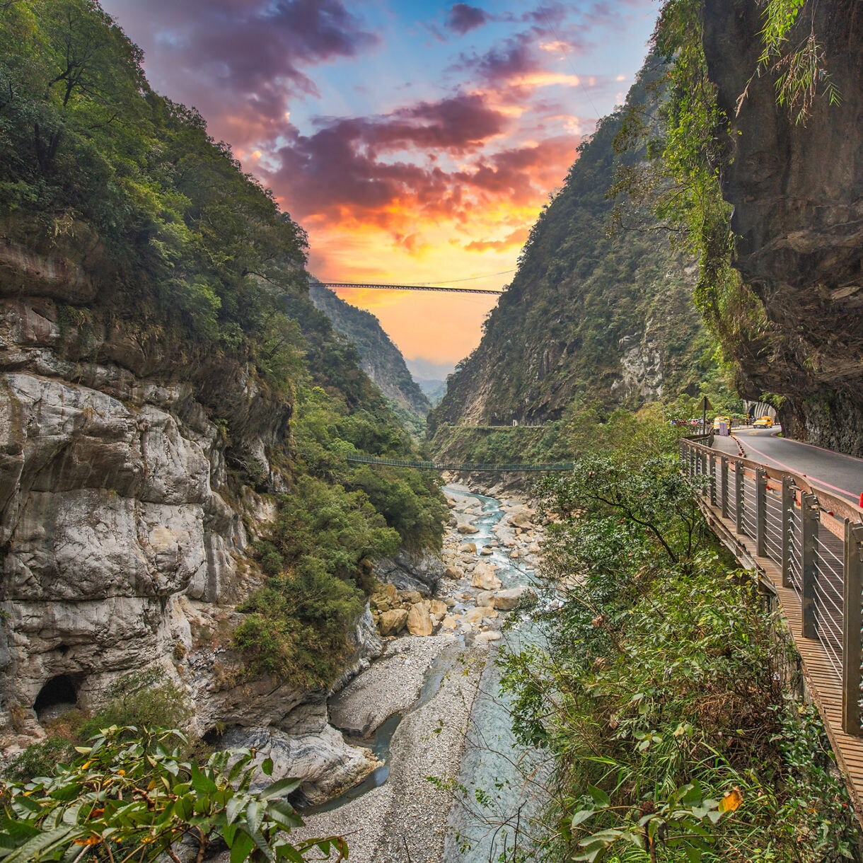 A dramatic view of Taroko Gorge in Hualien, Taiwan, featuring steep marble cliffs, a narrow river below, lush greenery and a walkway built along the rock face. A vibrant sunset glows between the cliffs, with suspension bridges visible in the distance.