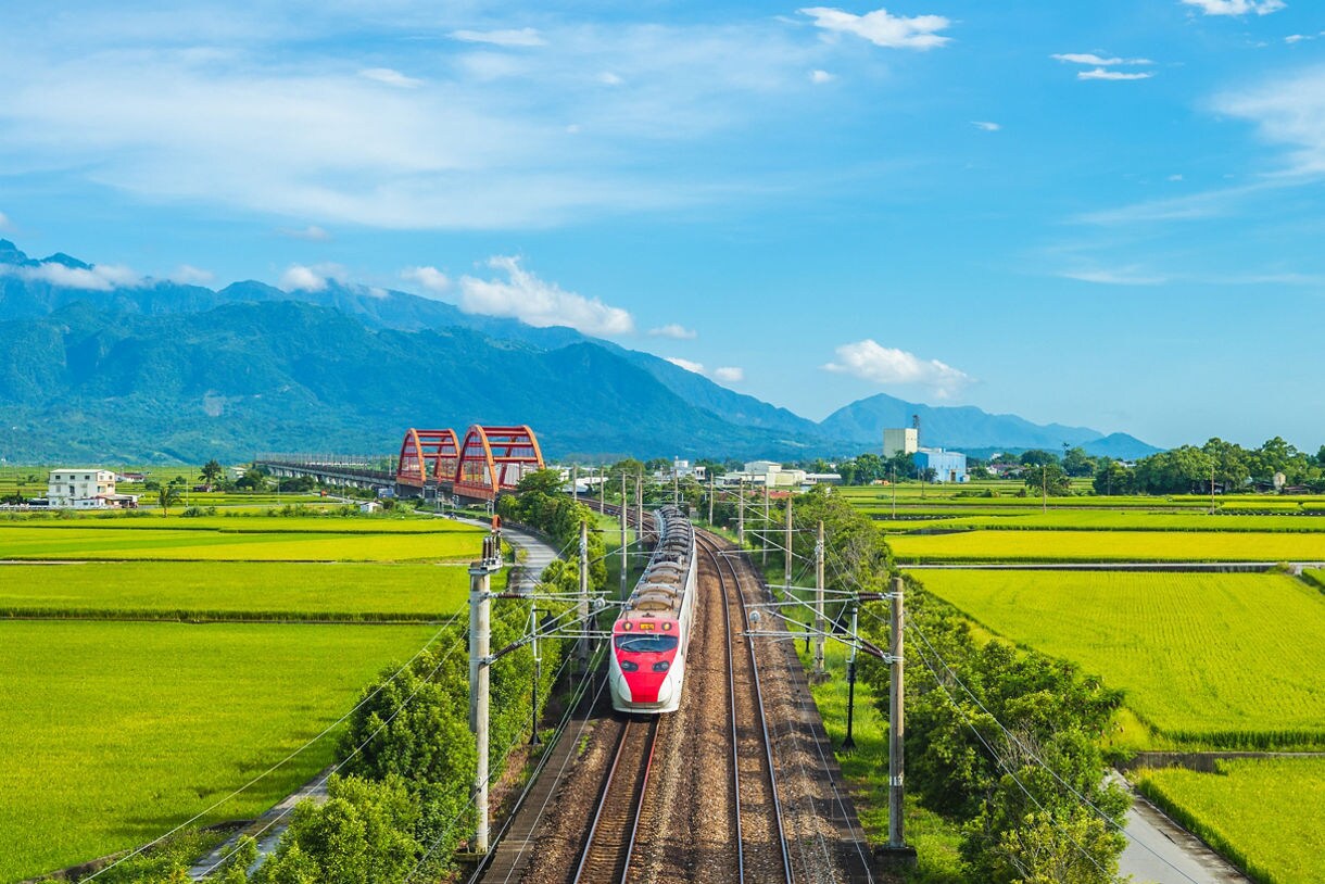 A red and white train crossing a bridge through green and yellow rice fields in Hualien, Taiwan, with blue mountains in the distance under a clear sky.