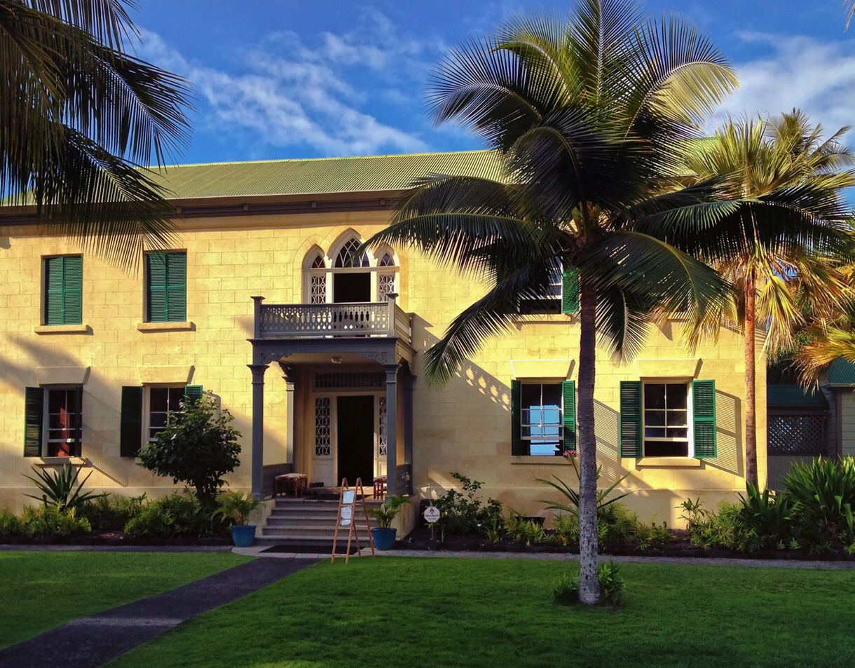Historic two-story Huliheʻe Palace with green shutters and palm trees in front, glowing in warm afternoon light.