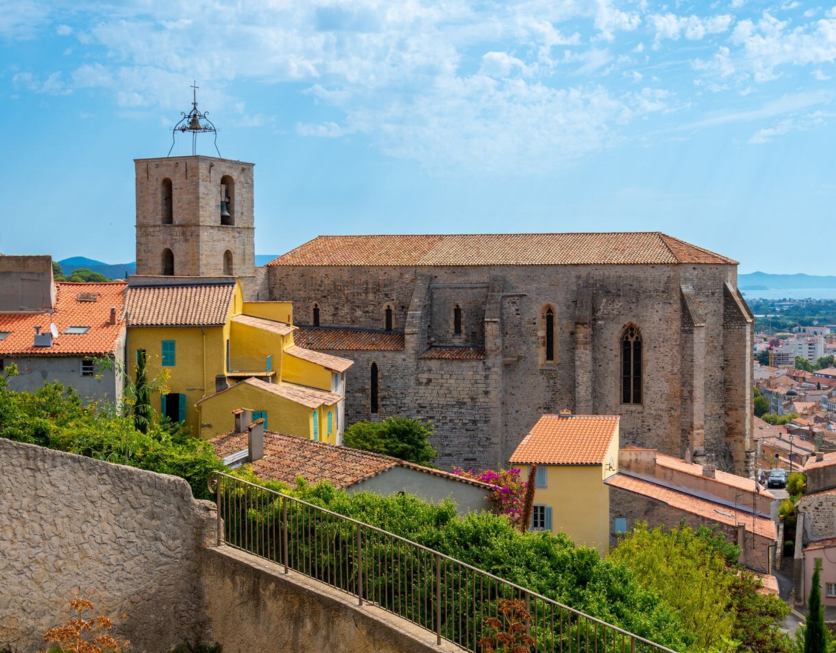 A stone medieval church in Hyères stands above a hillside of pastel yellow and orange houses with terracotta roofs, surrounded by greenery and overlooking a distant coastline under a bright blue sky.
