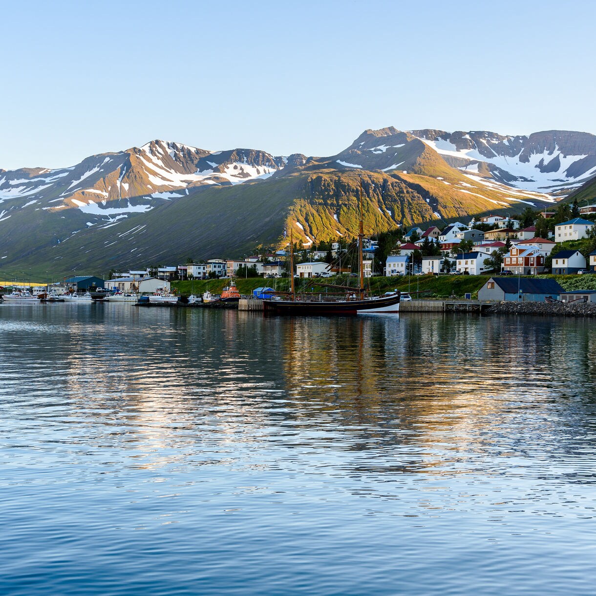 Calm fjord reflecting a small coastal village with bright houses and snow-dusted mountains in soft evening light.