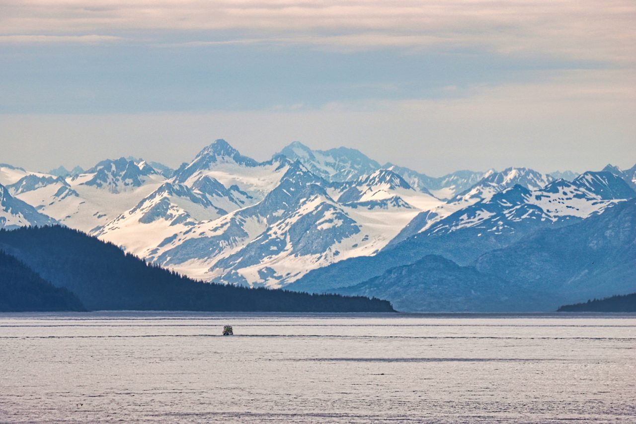 A small cruise ship crosses the calm gray waters of Icy Strait in Alaska, dwarfed by layers of forested coastline and massive snow-capped mountains under a pale overcast sky.