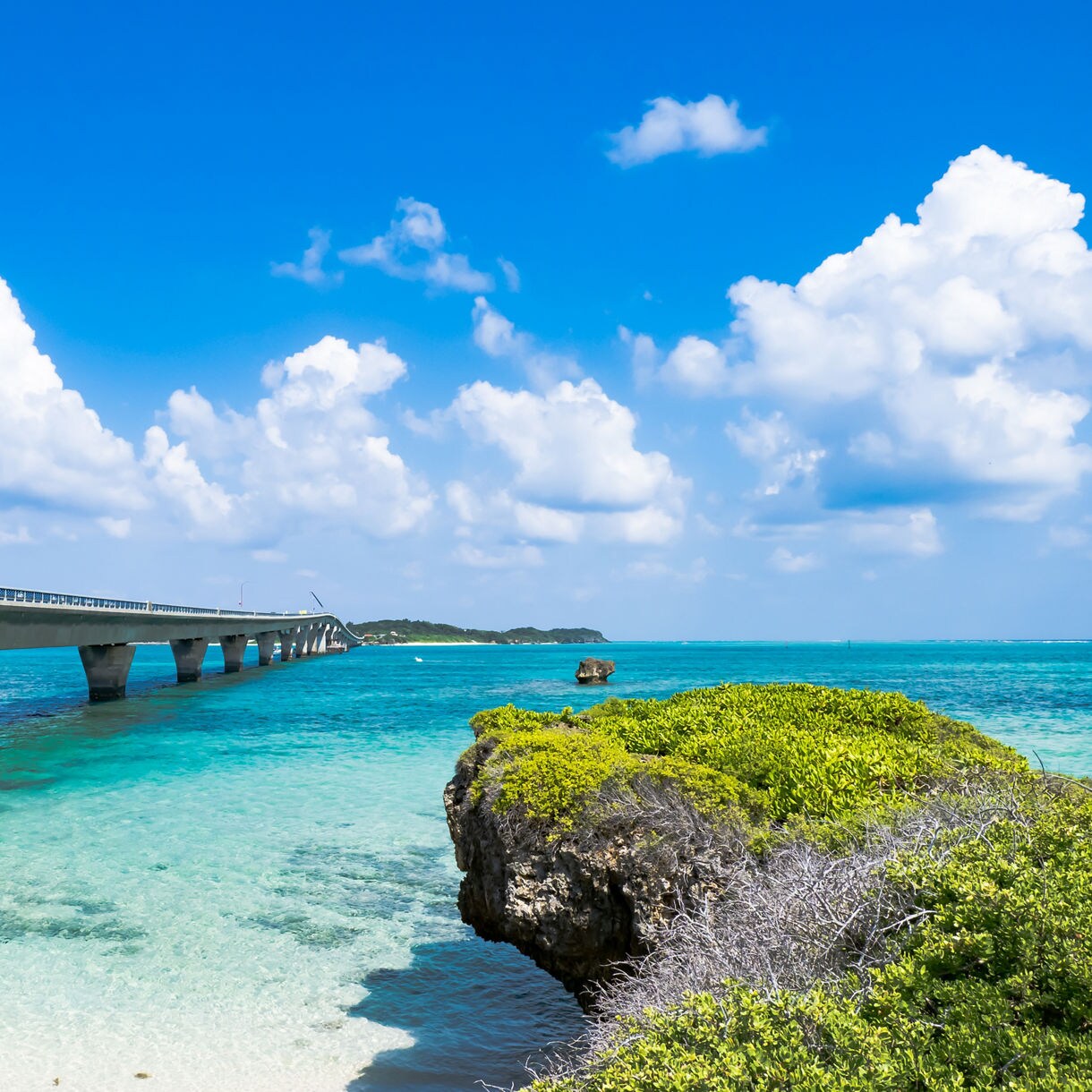Wide view of Ikema Bridge in Okinawa arching over bright turquoise water beside rocky greenery under a vivid blue sky with scattered clouds.
