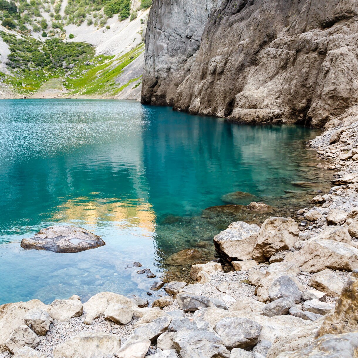 Clear blue lake surrounded by steep rocky cliffs and sparse green vegetation, with stones lining the shore in the foreground.