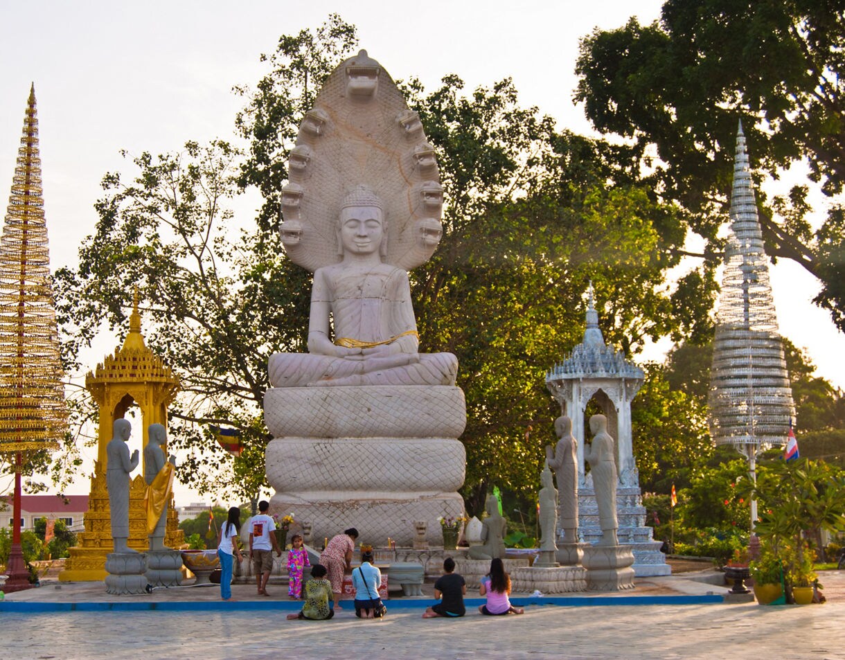 Large seated Buddha statue with a multi-headed naga behind it surrounded by smaller statues and people praying in an open courtyard.