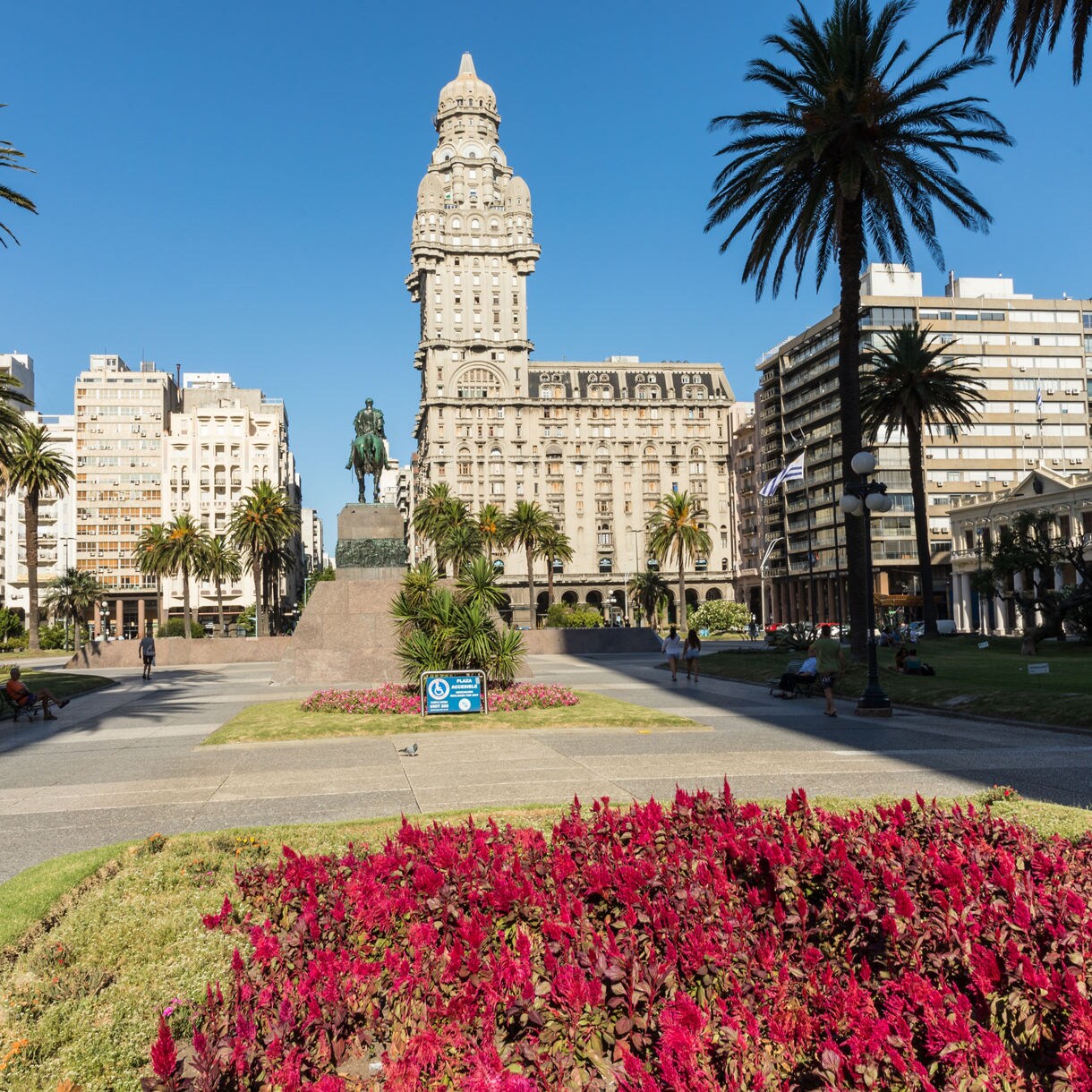 View of Montevideo’s Independence Square with red flowerbeds, tall palm trees, the central equestrian statue and the Palacio Salvo rising behind it.