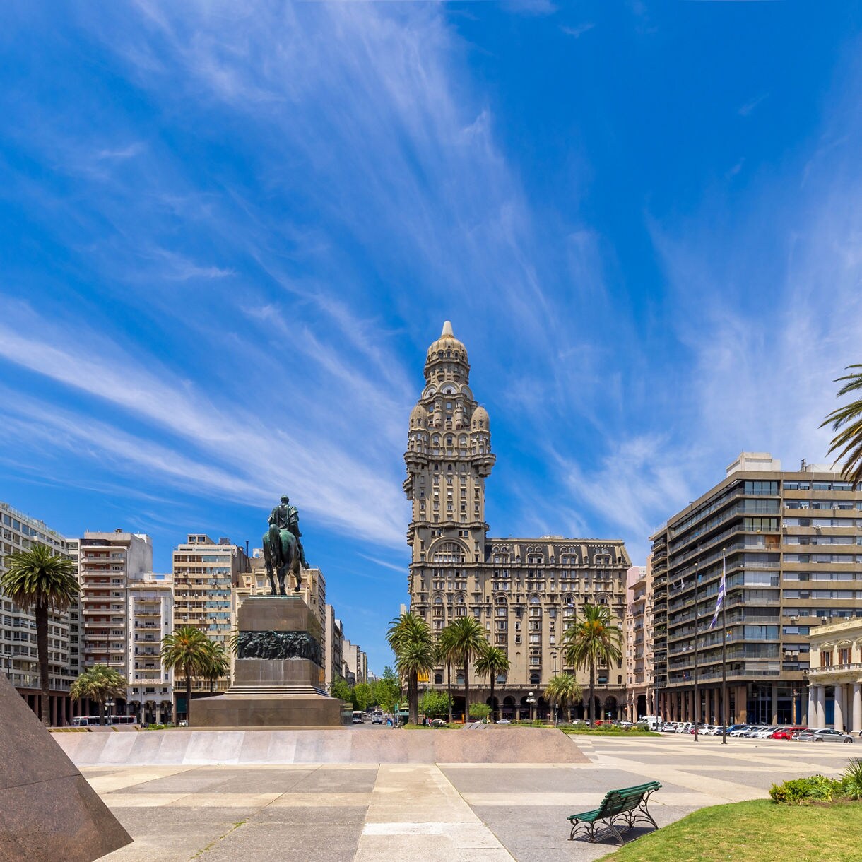 Expansive view of Independence Square in Montevideo featuring the equestrian statue of Artigas, tall palm trees and the ornate Palacio Salvo set against dramatic wispy clouds.