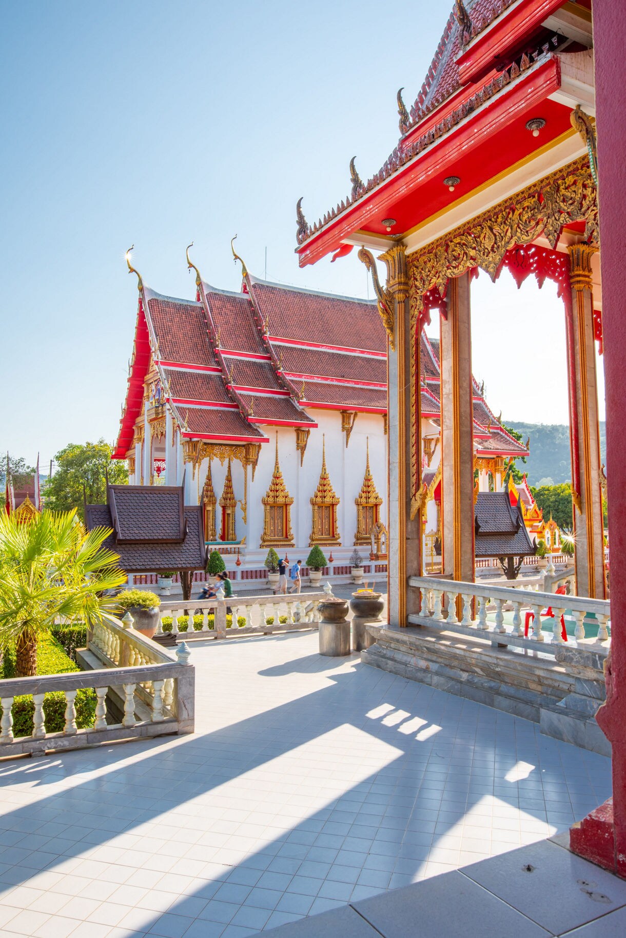 View of Wat Chalong in Phuket with red and gold roofs, intricate carvings and bright sunlight casting long shadows.