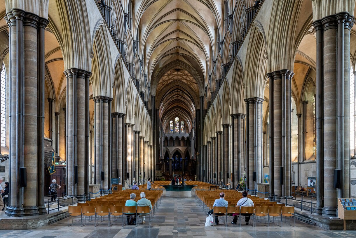 Interior view of Salisbury Cathedral’s nave, with tall Gothic columns, vaulted ceilings and rows of wooden chairs leading toward a stained glass window at the far end.