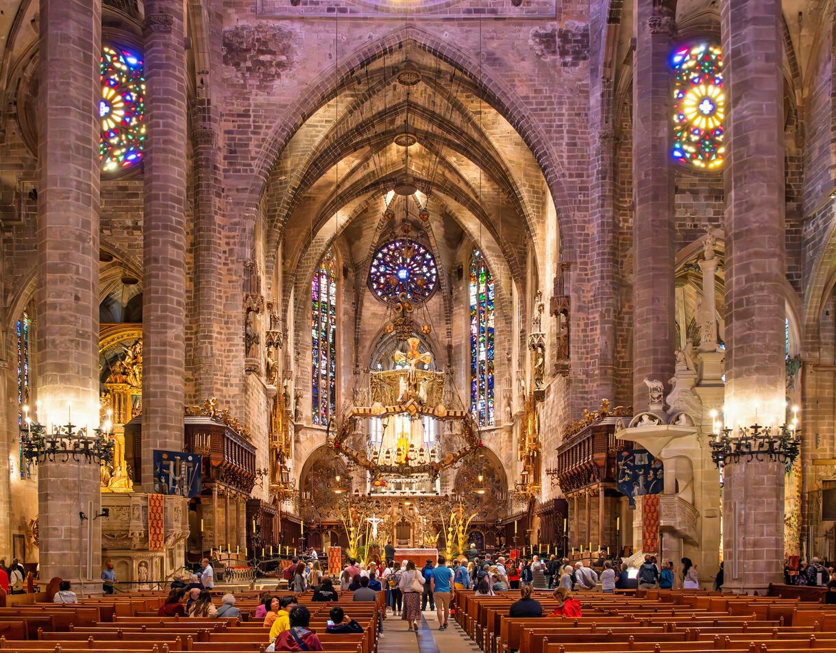 Interior view of La Seu Cathedral in Palma, Mallorca, featuring tall Gothic arches, intricate stained-glass windows and visitors admiring the ornate altar.
