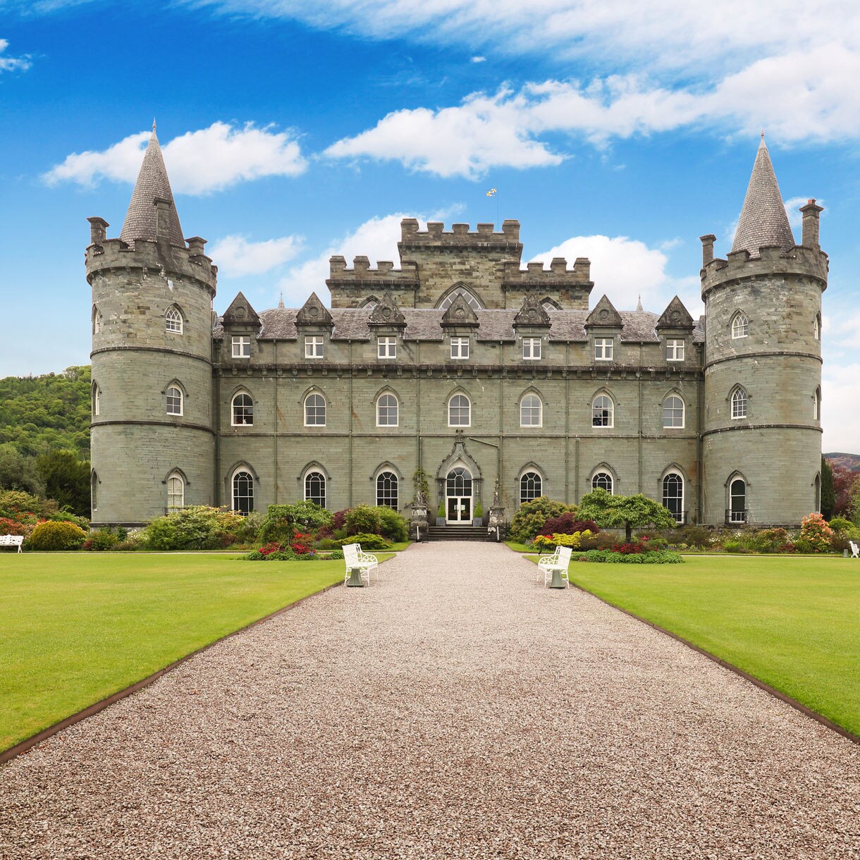 Inveraray Castle in Scotland, a grand stone building with turrets and battlements, fronted by a wide gravel path and lush green lawns surrounded by colorful shrubs.