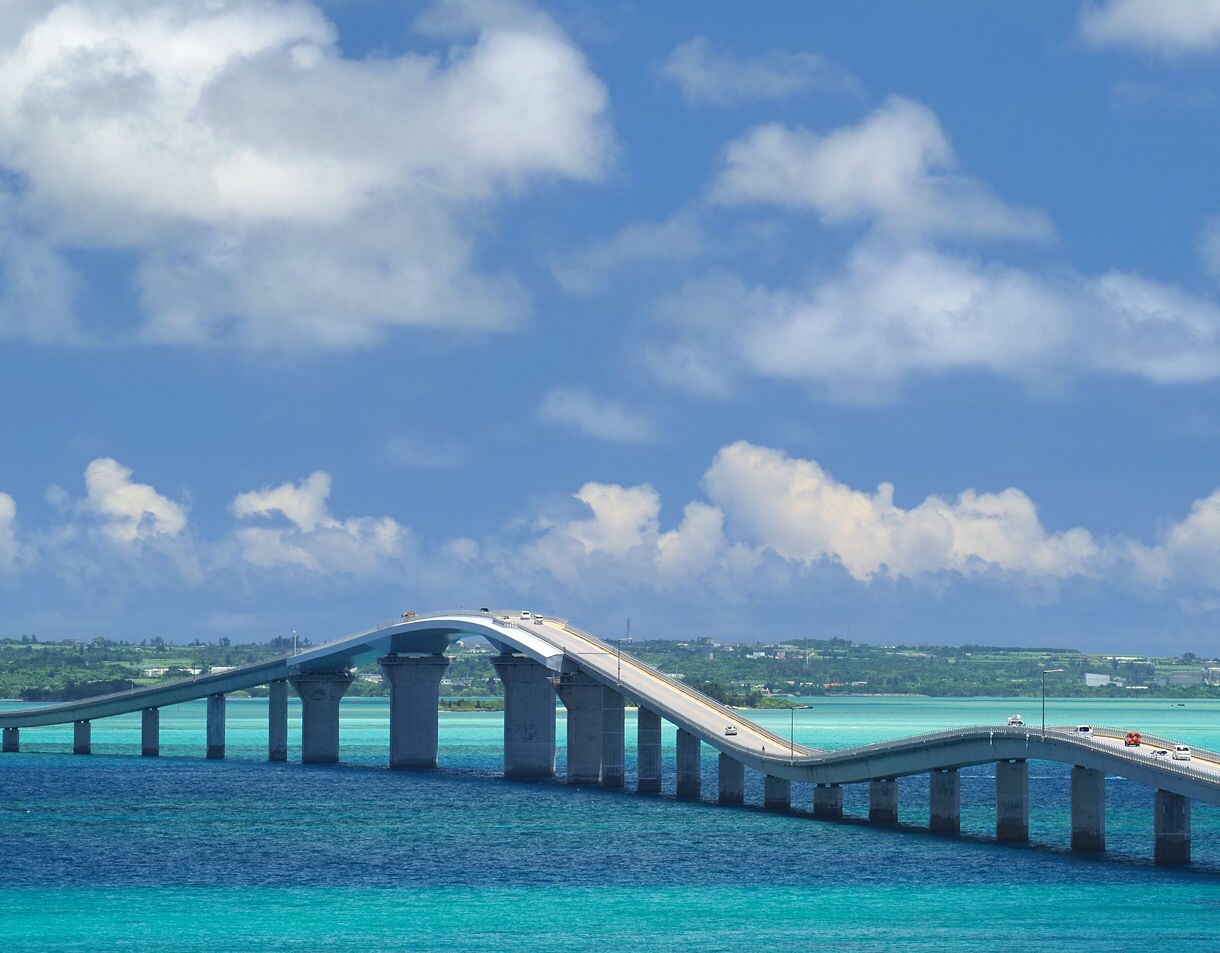 Long, gently undulating Irabu Ohashi Bridge stretching over bright turquoise and deep blue water with cars crossing and a boat nearby under a sky filled with soft white clouds.