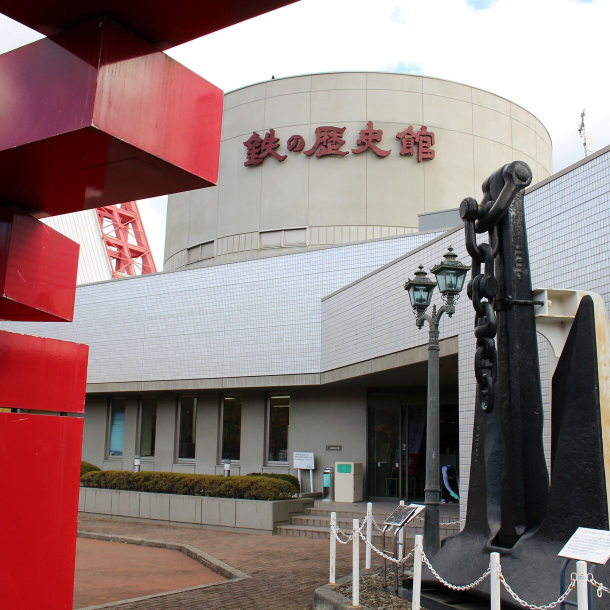Exterior view of the Iron and Steel Museum in Japan, featuring a large black ship anchor displayed near the entrance, red modernist metal sculptures in the foreground and a white tiled building with Japanese signage in the background.