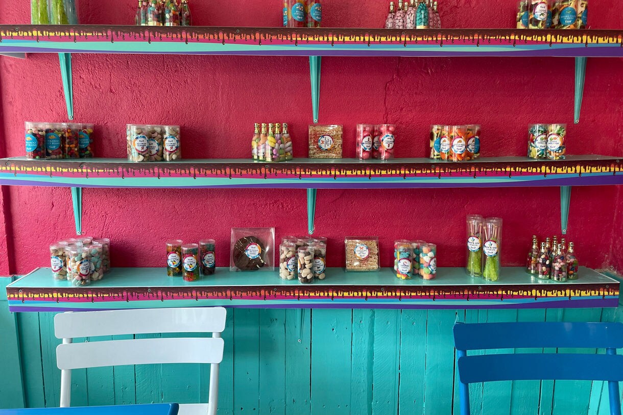 Display of candies and treats in jars and bottles arranged on turquoise shelves against a pink wall, with bright chairs in the foreground.
