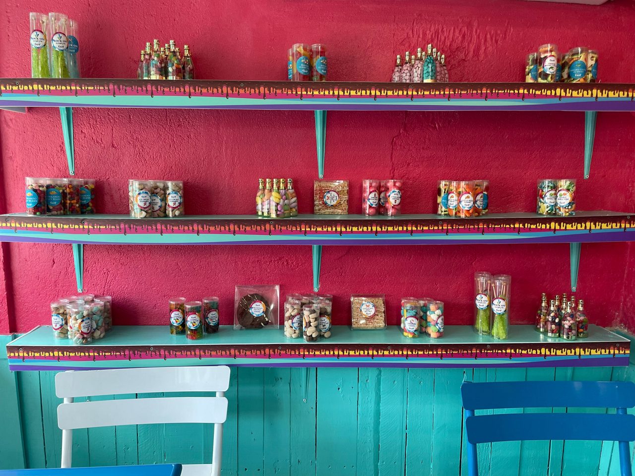 Display of candies and treats in jars and bottles arranged on turquoise shelves against a pink wall, with bright chairs in the foreground.