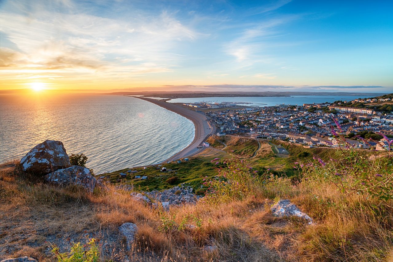 Elevated view of Chesil Beach and the town of Portland in Dorset at sunset, with the long shingle spit curving along the coast and warm sunlight reflecting on the sea.