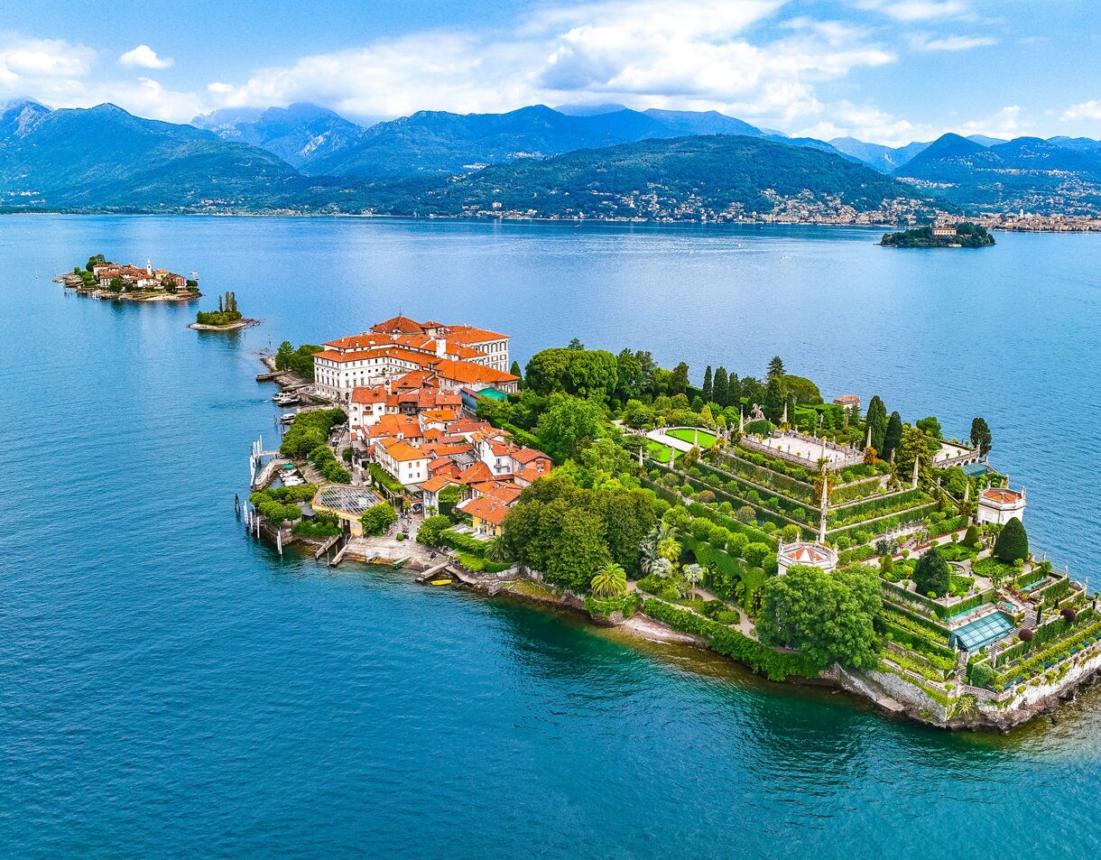 Aerial view of Isola Bella on Lake Maggiore, showing terraced Baroque gardens, red-roofed buildings and deep blue water surrounded by distant mountains.