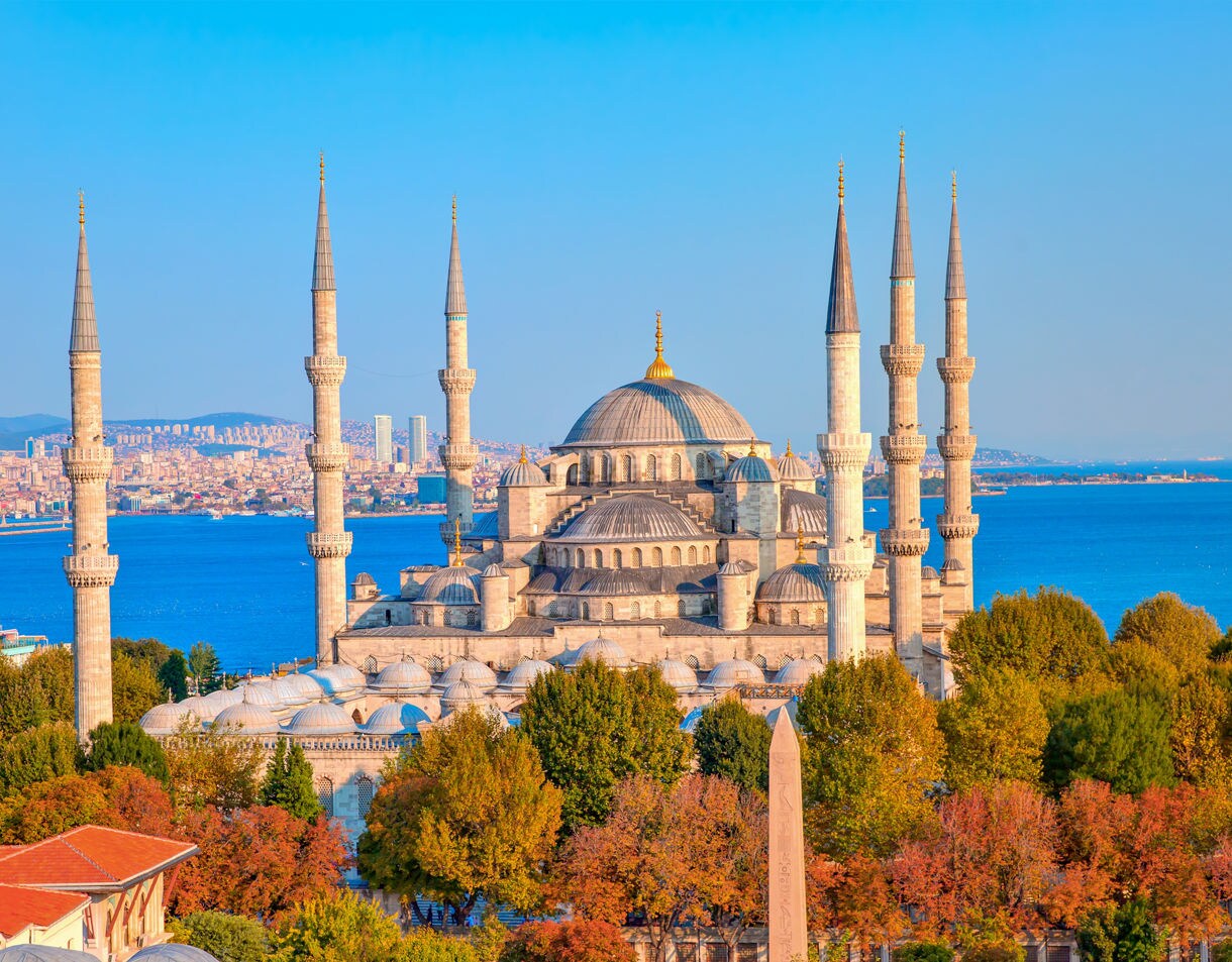 The Blue Mosque in Istanbul with its domes and six tall minarets surrounded by trees, set against the backdrop of the Bosphorus and city skyline.