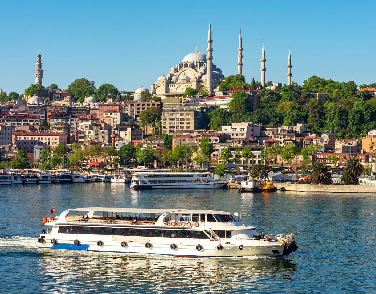  A boat cruising the Bosphorus in Istanbul with the Suleymaniye Mosque and its tall minarets rising above the city’s hillside buildings.