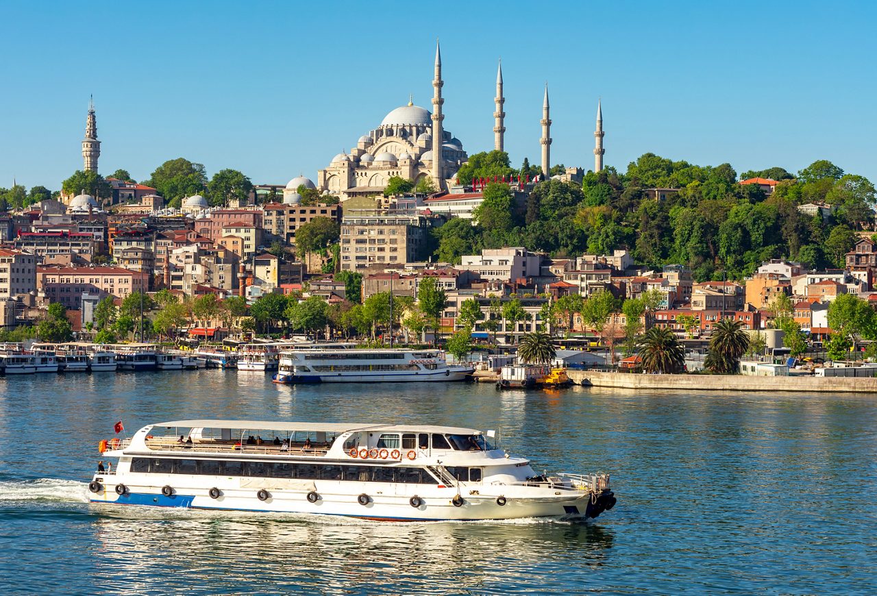 A white passenger boat gliding along the Bosphorus with Istanbul’s hillside cityscape behind it, featuring domed mosques, slender minarets, green trees and waterfront buildings under a clear blue sky.