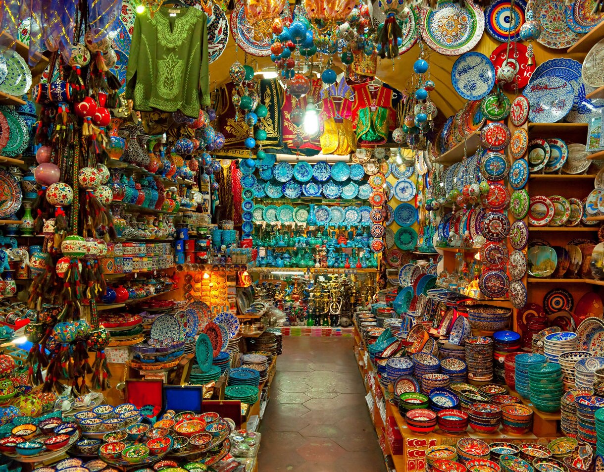 A vibrant stall in Istanbul’s Grand Bazaar filled with hand-painted ceramic plates, bowls, hanging lanterns and traditional textiles in bold colors.