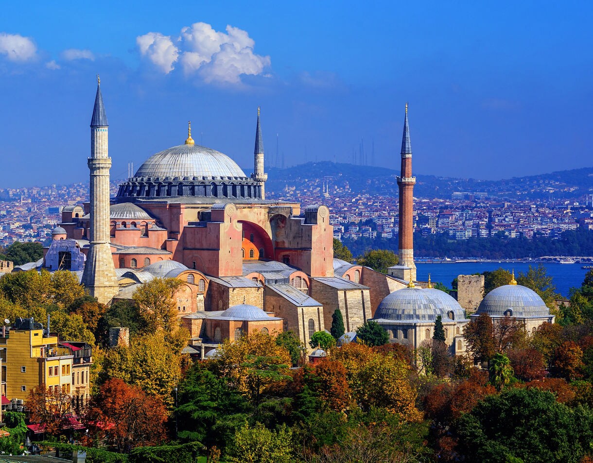 The Hagia Sophia in Istanbul with its massive dome and four slender minarets, surrounded by trees and colorful city buildings near the Bosphorus.
