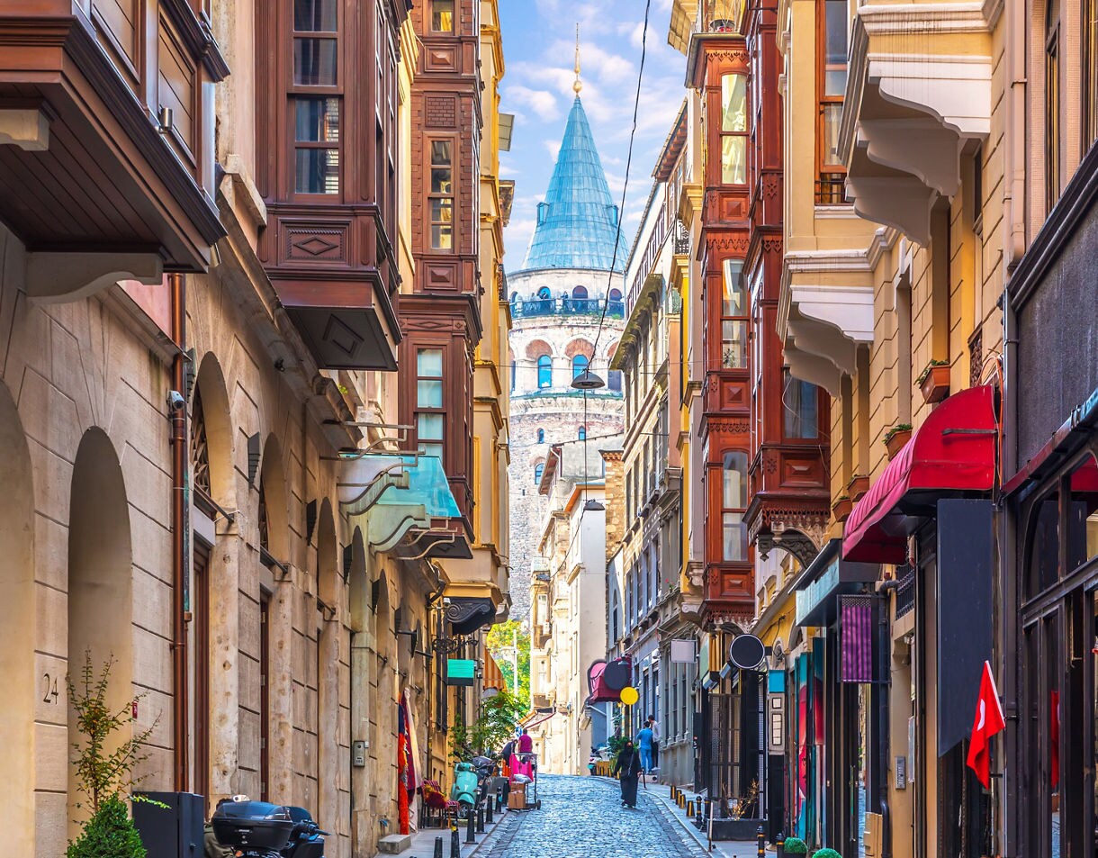 Narrow cobblestone street in Istanbul lined with colorful buildings and shops, leading to the historic Galata Tower in the distance.