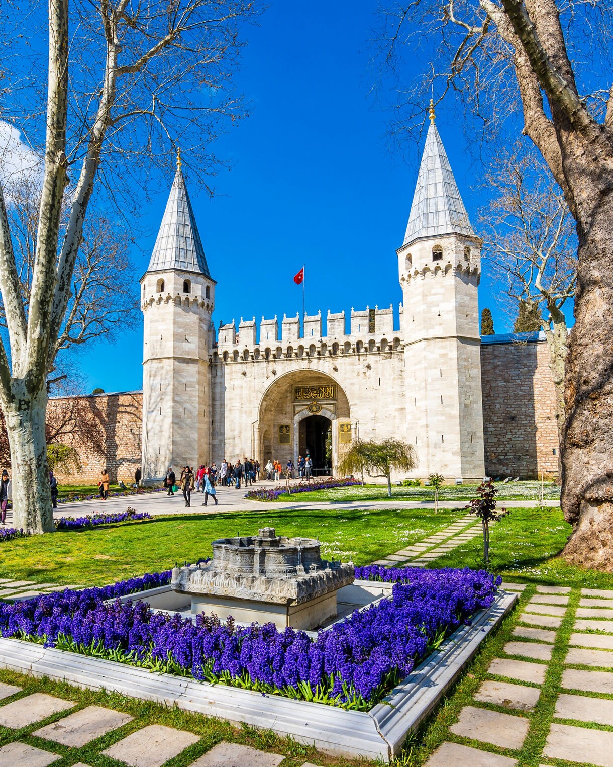 Entrance to Topkapi Palace in Istanbul with tall towers, stone walls, spring flowers and visitors walking through the gardens.